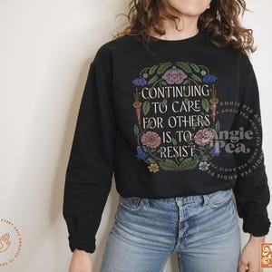 May include: Black sweatshirt with a floral design and the text "Continuing to care for others is to resist".