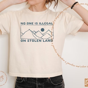 May include: A white t-shirt with a blue graphic design that says "No one is illegal on stolen land". The graphic features a mountain range with a sun in the background.