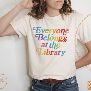 May include: A white t-shirt with the text "Everyone Belongs at the Library" in rainbow colors. The text is in a bold, playful font.