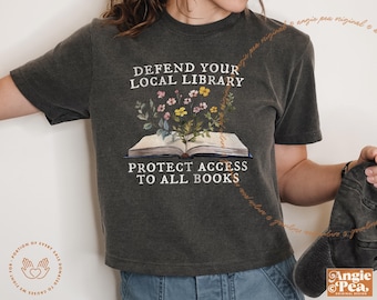 Op de afbeelding: Een donkergrijs T-shirt met de tekst "DEFEND YOUR LOCAL LIBRARY" en "PROTECT ACCESS TO ALL BOOKS" erop gedrukt. Het shirt heeft een bloemige illustratie van een boek met bloemen.