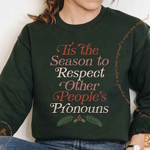 May include: A dark green sweatshirt with the text "Tis the Season to Respect Other People's Pronouns" in a festive font with red and white accents. The text is surrounded by a green leafy design with red berries.