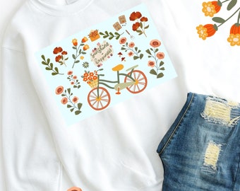 Op de afbeelding: Witte trui met een blauwe en witte afbeelding van een fiets met bloemen en de tekst "Sunshine Happiness". De trui ligt op een wit oppervlak met een paar blauwe jeans met distressed details.