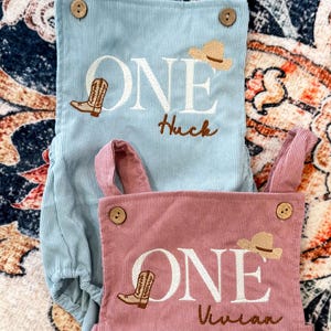 May include: Two corduroy baby rompers, one light blue and one dusty rose. Each features the word "ONE" in white, a cowboy boot and hat design, and a name. The light blue romper says "Huck", and the dusty rose romper says "Vivian".