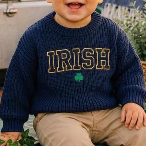 May include: A navy blue long-sleeve knit jumper featuring the word IRISH in yellow block letters with a green shamrock icon below the text. The jumper has a ribbed crew neckline, cuffs, and hem, paired with tan trousers and white trainers.