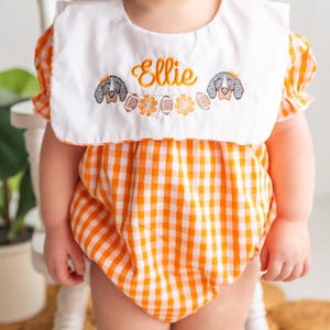 Puede incluir: Un pelele de bebé a cuadros naranja y blanco con un babero blanco. El babero está bordado con el nombre "Ellie", dos ilustraciones de perros y diseños florales. El pelele tiene mangas cortas abullonadas.