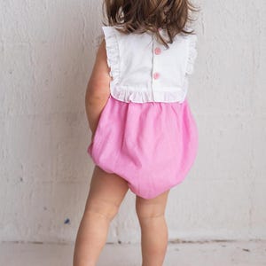 May include: A baby's romper with a white top and pink bottom. The top has ruffled sleeves and three pink buttons down the back. The bottom is a bubble style with vertical stripes. The baby is facing away from the camera.
