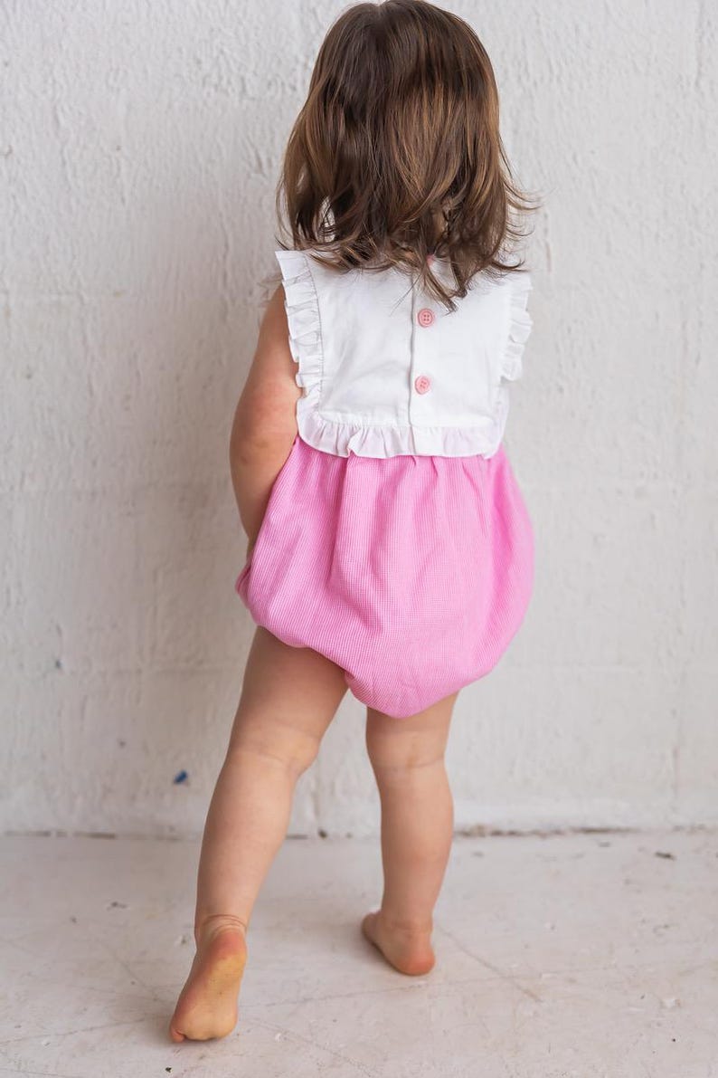 May include: A baby's romper with a white top and pink bottom. The top has ruffled sleeves and three pink buttons down the back. The bottom is a bubble style with vertical stripes. The baby is facing away from the camera.