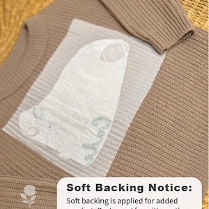 May include: A tan knitted sweater with a ribbed design. A white, textured backing is applied for comfort. The image includes the text "Soft Backing Notice" and care instructions for gentle washing and air drying.