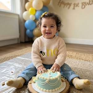 May include: A child's first birthday celebration. The child wears a beige sweater with a bee and the word "one" in yellow. A light blue cake sits on a wooden slice. Balloons in yellow, white, and blue decorate the background.