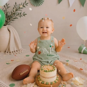 May include: A sage green romper featuring the embroidered text ONE with a football graphic and a name, worn by a child sitting on a textured mat next to a small cake and a brown football, surrounded by green and white balloons and confetti.