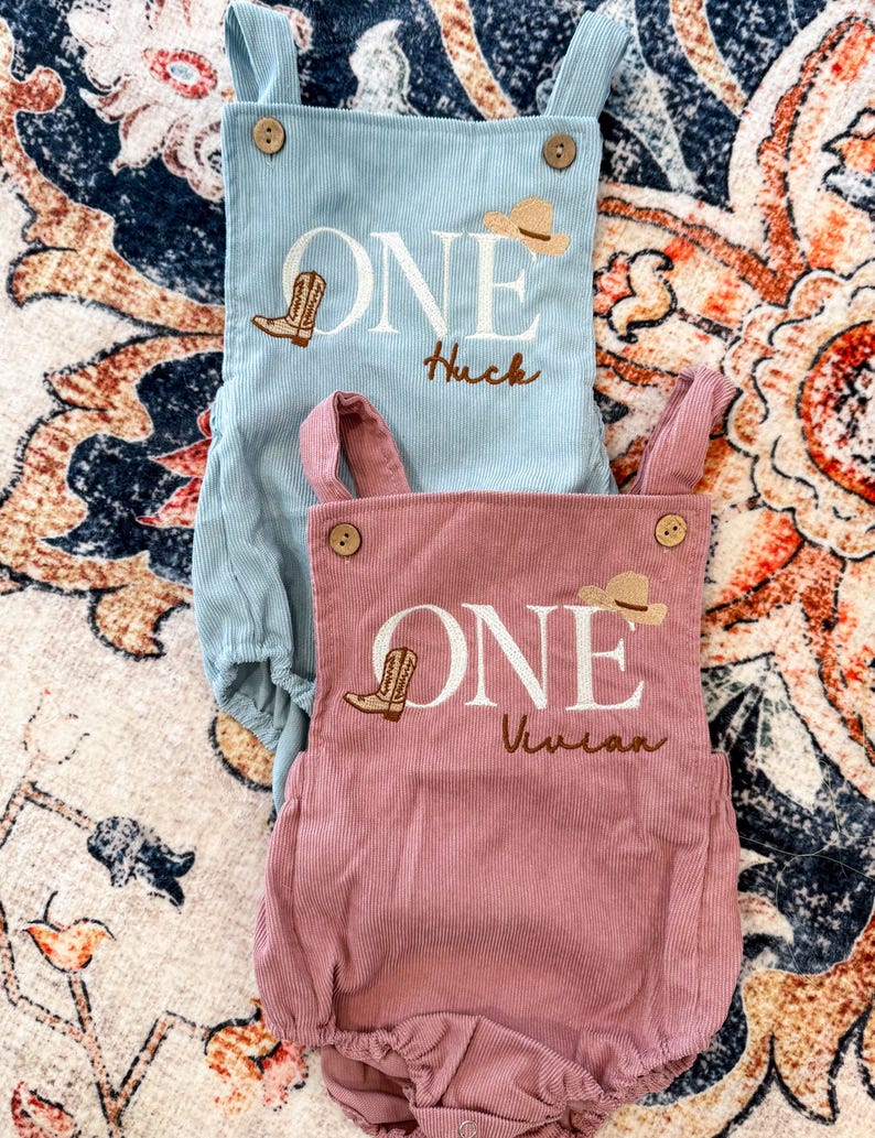 May include: Two corduroy baby rompers, one light blue and one dusty rose. Each features the word "ONE" with a cowboy boot and hat design, and a name embroidered below. Wooden button details.