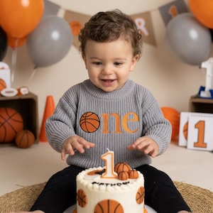 May include: A child wearing a grey sweater with an orange basketball and the word "one" embroidered on it. The child is in front of a birthday cake decorated with basketballs and a number one candle. Balloons and basketball decorations are in the background.