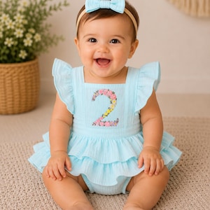 May include: A smiling baby wearing a light blue romper with a floral embroidered number 2. The romper has ruffled sleeves and a tiered skirt. A matching bow headband completes the outfit. The baby is sitting on a neutral-colored rug.