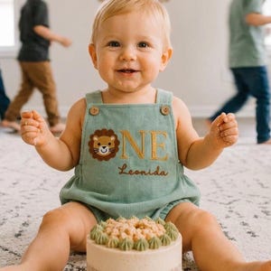 May include: A sage green corduroy romper featuring a brown embroidered lion face, the gold stitched text ONE, and the name Leonida in brown cursive script. The garment has two wooden buttons on the front straps and a gathered elastic waist.
