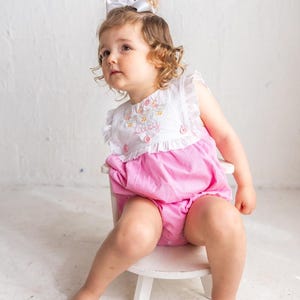 May include: A little girl wearing a white and pink seersucker romper with the name "Lucy" embroidered on the front. The romper has ruffle sleeves and is paired with a white bow. She is sitting on a small white chair.