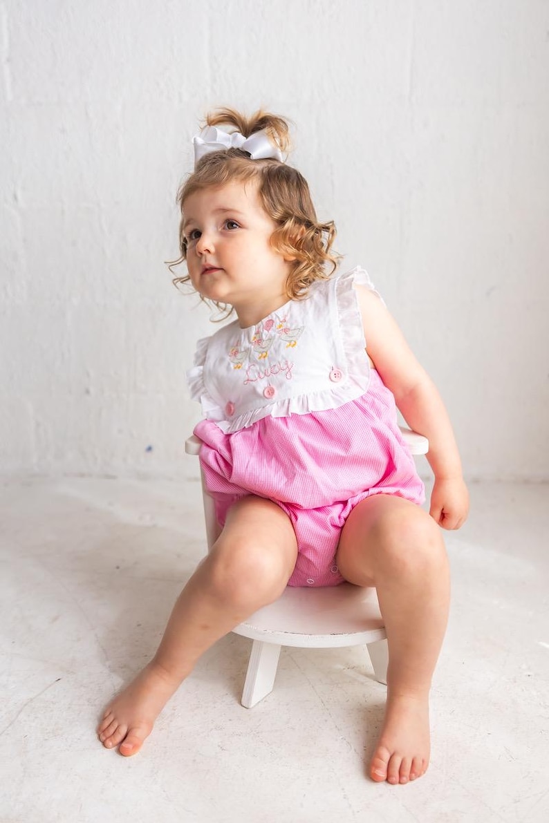 May include: A little girl wearing a white and pink seersucker romper with the name "Lucy" embroidered on the front. The romper has ruffle sleeves and is paired with a white bow. She is sitting on a small white chair.