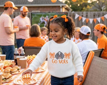 May include: A young girl wearing a cream-colored sweater with a dog graphic and the text "Go Vols" smiles at an outdoor gathering. The sweater has orange bows on each side of the dog graphic. The table is covered with an orange and white checkered tablecloth.