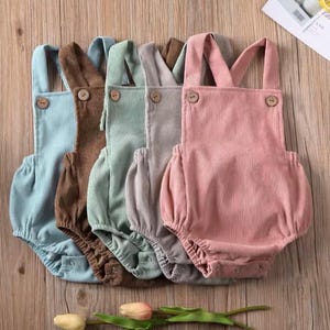 May include: Five pastel-coloured corduroy baby rompers: light blue, brown, green, grey, and pink. Each has wooden buttons on the straps and a gathered bottom. The rompers are arranged on a wooden surface.