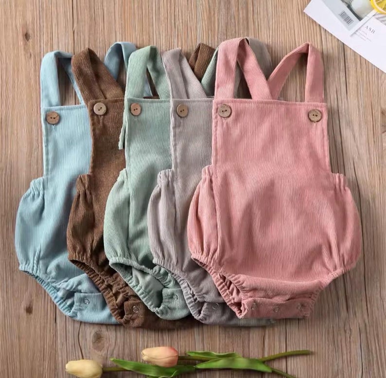 May include: Five pastel-coloured corduroy baby rompers: light blue, brown, green, grey, and pink. Each has wooden buttons on the straps and a gathered bottom. The rompers are arranged on a wooden surface.