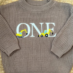 Peut inclure: Pull en tricot marron avec le mot "ONE" en blanc. Le "O" est orné d'une excavatrice jaune et noire, et le "E" d'un bulldozer jaune et noir. Le pull a un col rond côtelé et des poignets côtelés.