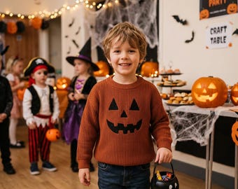 May include: A young child smiles, wearing a brown sweater with a black jack-o'-lantern face. The child holds a black candy bucket. Other children in costumes and Halloween decorations are in the background. A sign reads "Halloween Party."