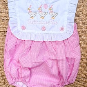 May include: A pink and white gingham baby bubble romper with a white bib-style top. The top features embroidered ducks wearing party hats and holding balloons, with the text "Birthday Girl". The romper has ruffle detailing.