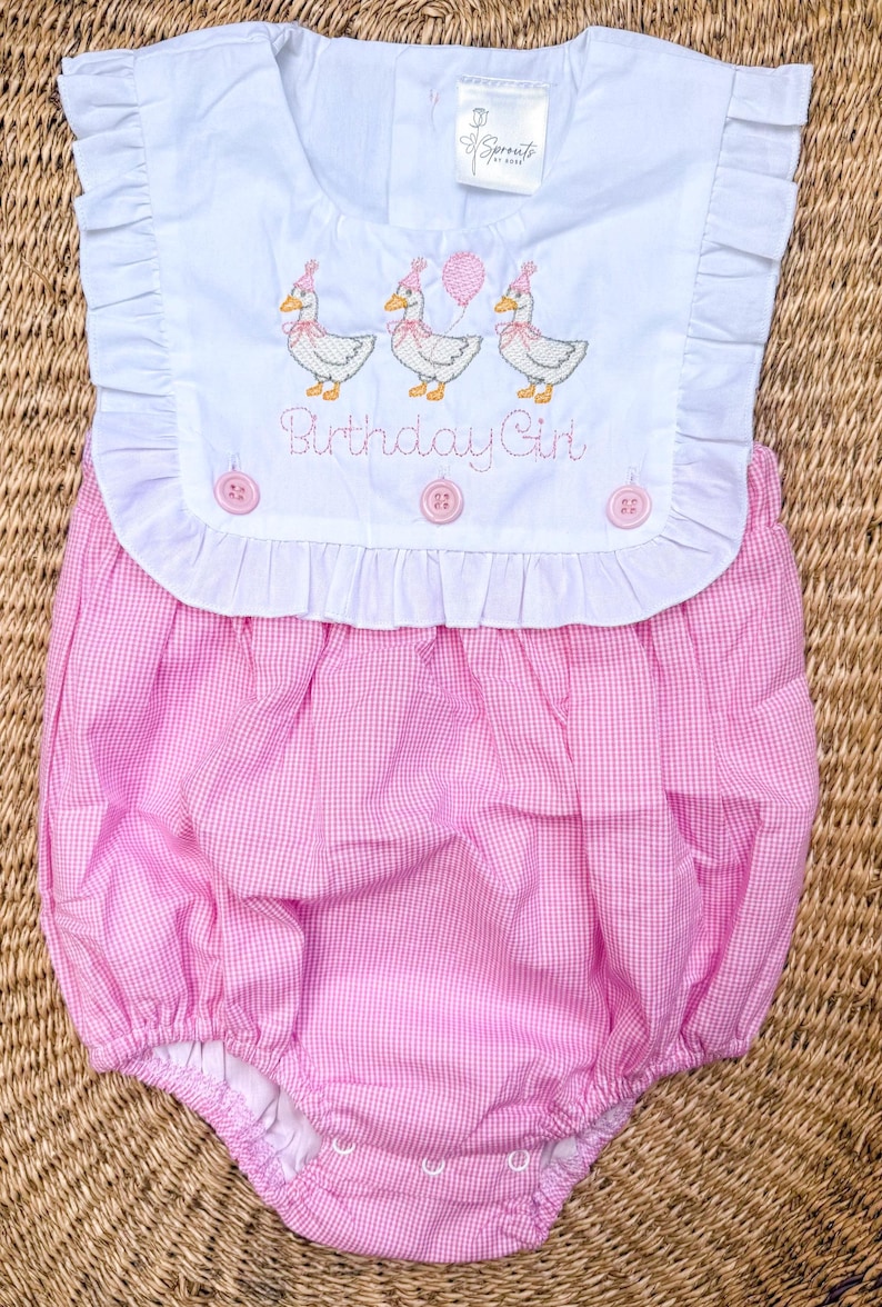 May include: A pink and white gingham baby bubble romper with a white bib-style top. The top features embroidered ducks wearing party hats and holding balloons, with the text "Birthday Girl". The romper has ruffle detailing.