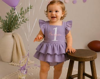 May include: A lavender-colored sleeveless romper with ruffled sleeves and a tiered skirt. The bodice features a white embroidered number one with the name Harper stitched in pink cursive script across the center of the number.
