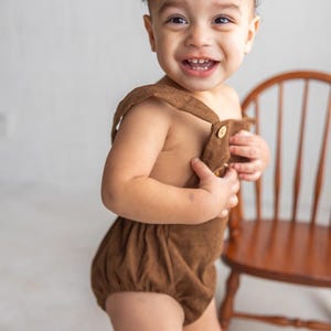 May include: A smiling toddler in a brown corduroy romper with button fastenings. The romper features suspender straps and a gathered bottom. A wooden chair is in the background.
