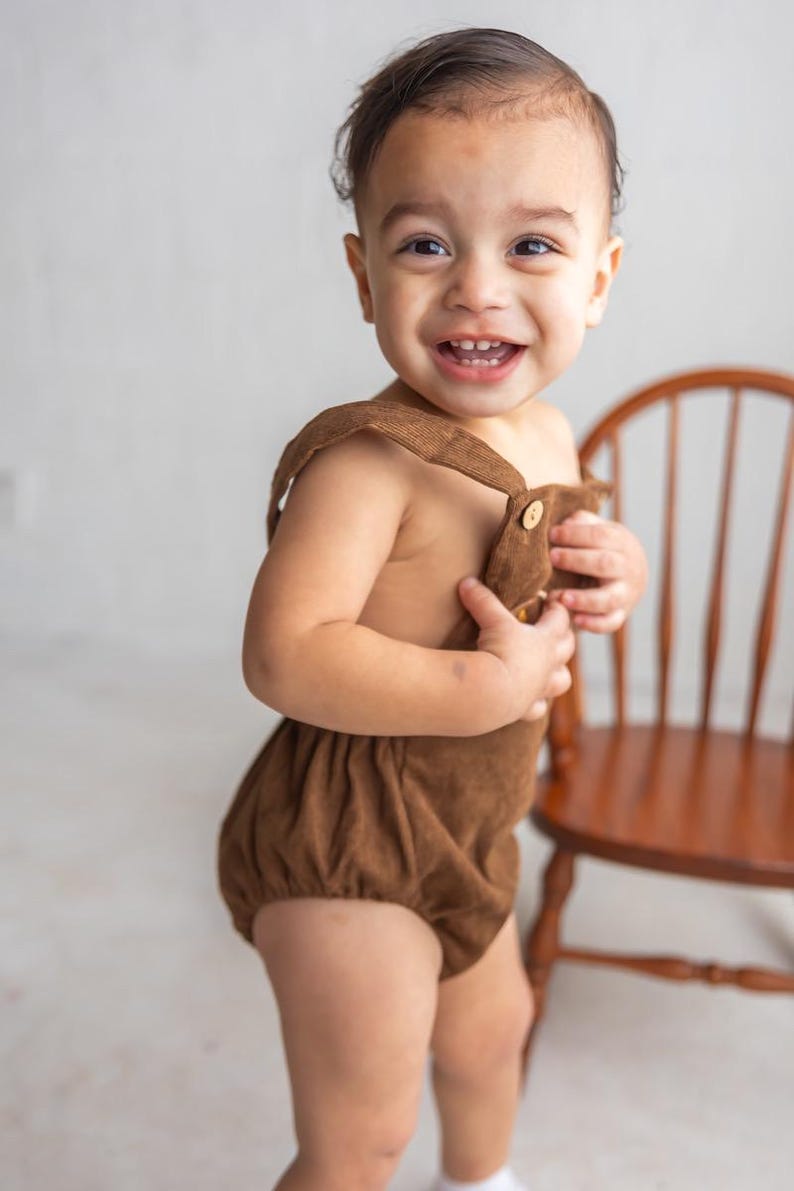 May include: A smiling toddler in a brown corduroy romper with button fastenings. The romper features suspender straps and a gathered bottom. A wooden chair is in the background.