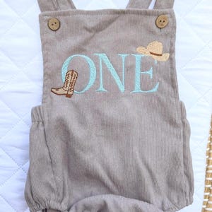 May include: A taupe corduroy romper with suspender straps and wooden buttons. The word "ONE" is embroidered in light blue, along with a cowboy boot and a cowboy hat. The romper is designed for a baby's first birthday.