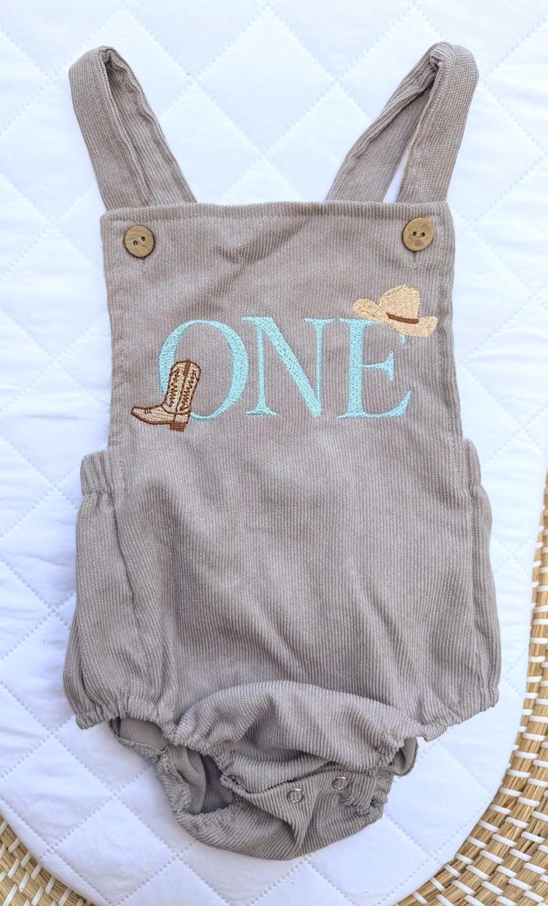 May include: A taupe corduroy romper with suspender straps and wooden buttons. The word "ONE" is embroidered in light blue, along with a cowboy boot and a cowboy hat. The romper is designed for a baby's first birthday.