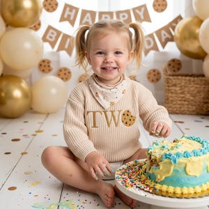May include: A young child with pigtails smiles, covered in frosting, celebrating a birthday. The child wears a beige sweater with "TWO" and a cookie graphic. A blue and yellow frosted cake sits nearby, surrounded by sprinkles. Balloons and a banner that says "HAPPY BIRTHDAY" are in the background.
