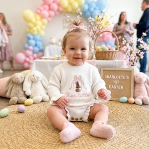 May include: A baby wearing a cream-colored knit romper with a bunny and bow embroidery. The romper has ruffle trim and wooden buttons. The text "Charlotte's 1st Easter" is displayed on a sign. Easter-themed decorations include balloons, stuffed bunnies, and Easter eggs.