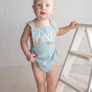 May include: A pale blue corduroy romper featuring the embroidered word "ONE", a cowboy boot, and a hat. The romper has buttoned straps and is worn by a baby. The baby stands near a small white ladder.