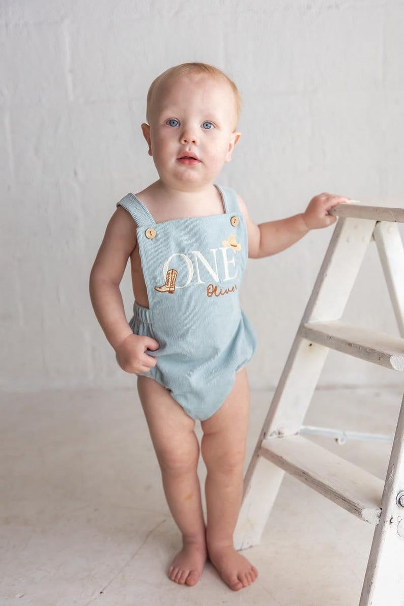 May include: A pale blue corduroy romper featuring the embroidered word "ONE", a cowboy boot, and a hat. The romper has buttoned straps and is worn by a baby. The baby stands near a small white ladder.