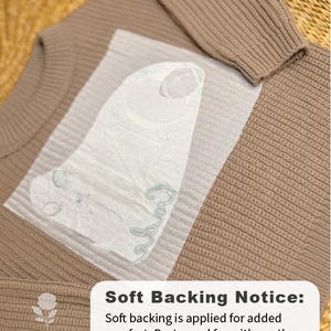 May include: A close-up of a tan sweater with a ribbed texture. A white, textured backing is applied to the sweater for added comfort. The text "Soft Backing Notice" is visible, along with care instructions.