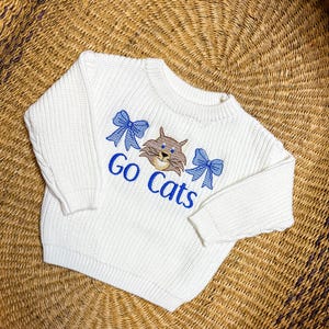 May include: Cream-colored sweater with the embroidered text "Go Cats". The design includes a cartoon cat face and two blue bows. The sweater is made of a thick knit material.