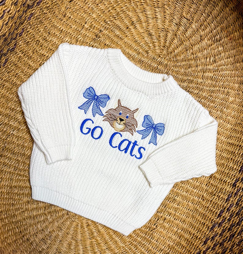 May include: Cream-colored sweater with the embroidered text "Go Cats". The design includes a cartoon cat face and two blue bows. The sweater is made of a thick knit material.