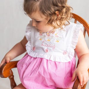 May include: A pink and white gingham romper with a white top featuring embroidered ducks and the name "Lucy". The romper has ruffled sleeves and is buttoned in the front. A white bow is in the child's hair.