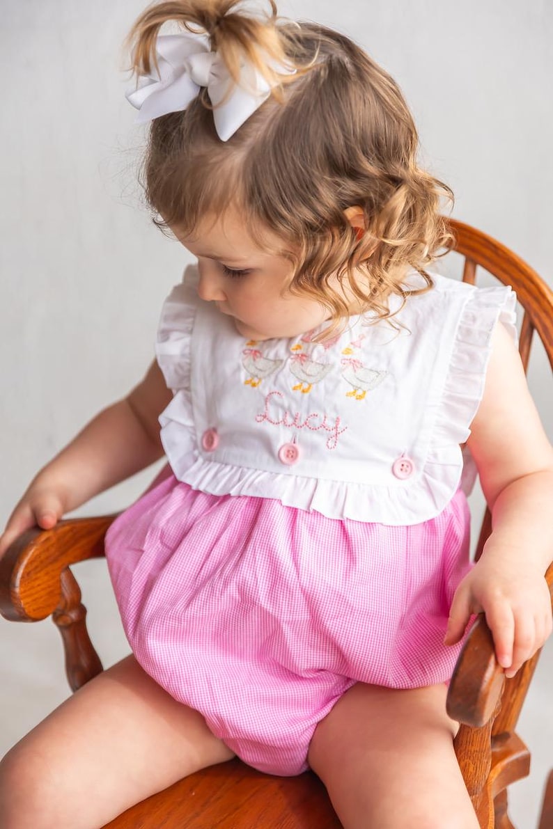 May include: A pink and white gingham romper with a white top featuring embroidered ducks and the name "Lucy". The romper has ruffled sleeves and is buttoned in the front. A white bow is in the child's hair.
