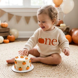 Puede incluir: Un bebé con un suéter color crema con una calabaza naranja y la palabra "one" en naranja. Un pastel pequeño decorado con calabazas está frente al bebé. Las calabazas y los globos están en el fondo.