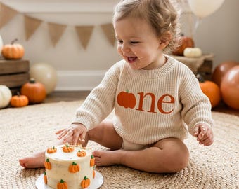 Op de afbeelding: Een baby draagt een crèmekleurige trui met een oranje pompoen en het woord "one" in oranje. Een kleine taart versierd met pompoenen staat voor de baby. Pompoenen en ballonnen zijn op de achtergrond.
