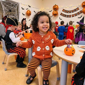 May include: A child smiles, wearing an orange cardigan with white ghost appliqués and matching striped leggings. The child holds a pumpkin-shaped candy bucket. The background features Halloween decorations, including balloons and a banner that says "Happy Halloween".