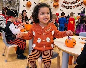 May include: A young child smiles, wearing an orange cardigan with white ghost appliqués and matching striped leggings. The child holds a pumpkin-shaped candy bucket. The background features Halloween decorations, including balloons and a banner that says "Happy Halloween".