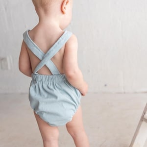May include: A pale blue baby romper with cross-over straps at the back. The romper features a gathered waist and a ribbed texture. The baby is facing away from the camera.