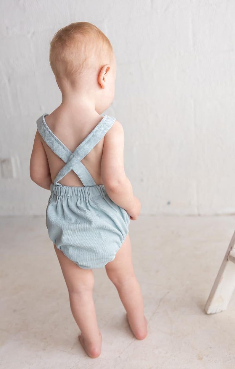 May include: A pale blue baby romper with cross-over straps at the back. The romper features a gathered waist and a ribbed texture. The baby is facing away from the camera.