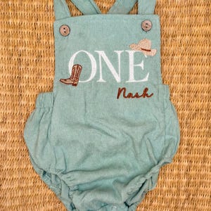 May include: A light teal corduroy baby romper with suspender straps and wooden buttons. The romper features the word "ONE" in white, a cowboy boot and hat, and the name "Nash" embroidered in brown. A cute outfit for a baby's first birthday.