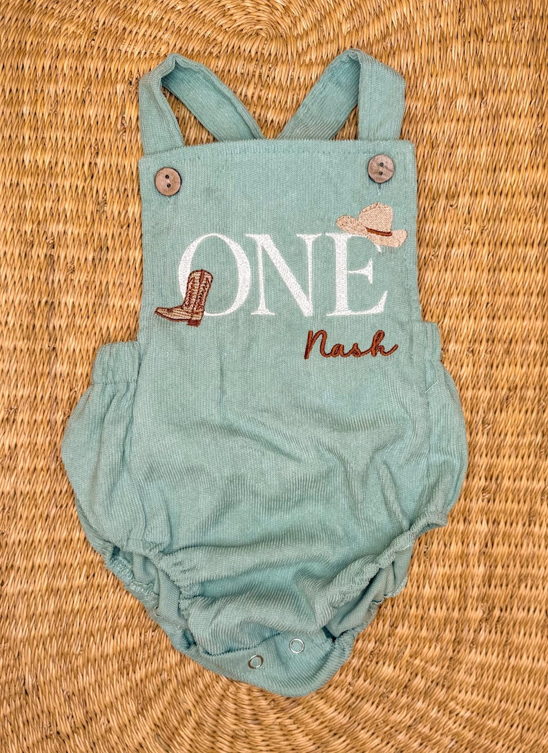 May include: A light teal corduroy baby romper with suspender straps and wooden buttons. The romper features the word "ONE" in white, a cowboy boot and hat, and the name "Nash" embroidered in brown. A cute outfit for a baby's first birthday.