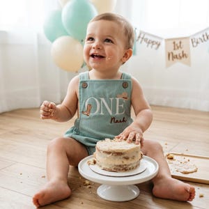 May include: A baby in a light green corduroy romper with "ONE" embroidered on the front, plus a cowboy boot and hat. The baby smiles in front of a small cake on a white stand. Balloons and a banner saying "Happy 1st Birthday Nash" are in the background. The cake is approximately 10 cm in diameter.
